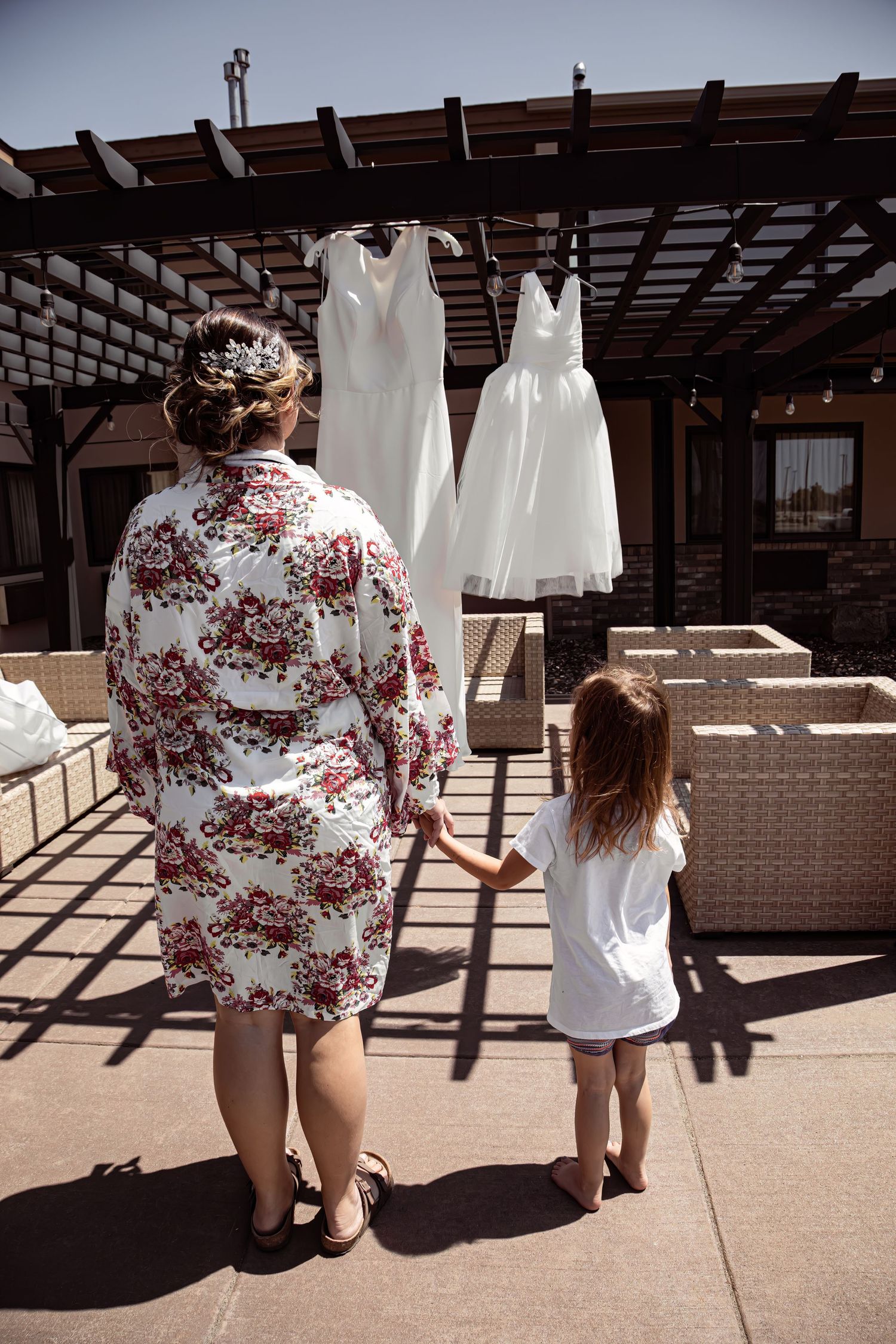 Two white wedding dresses hanging on outdoor pergola as two people look up at them.