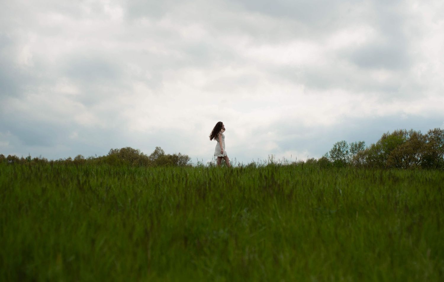 Distant figure stands alone in a green meadow under cloudy skies.