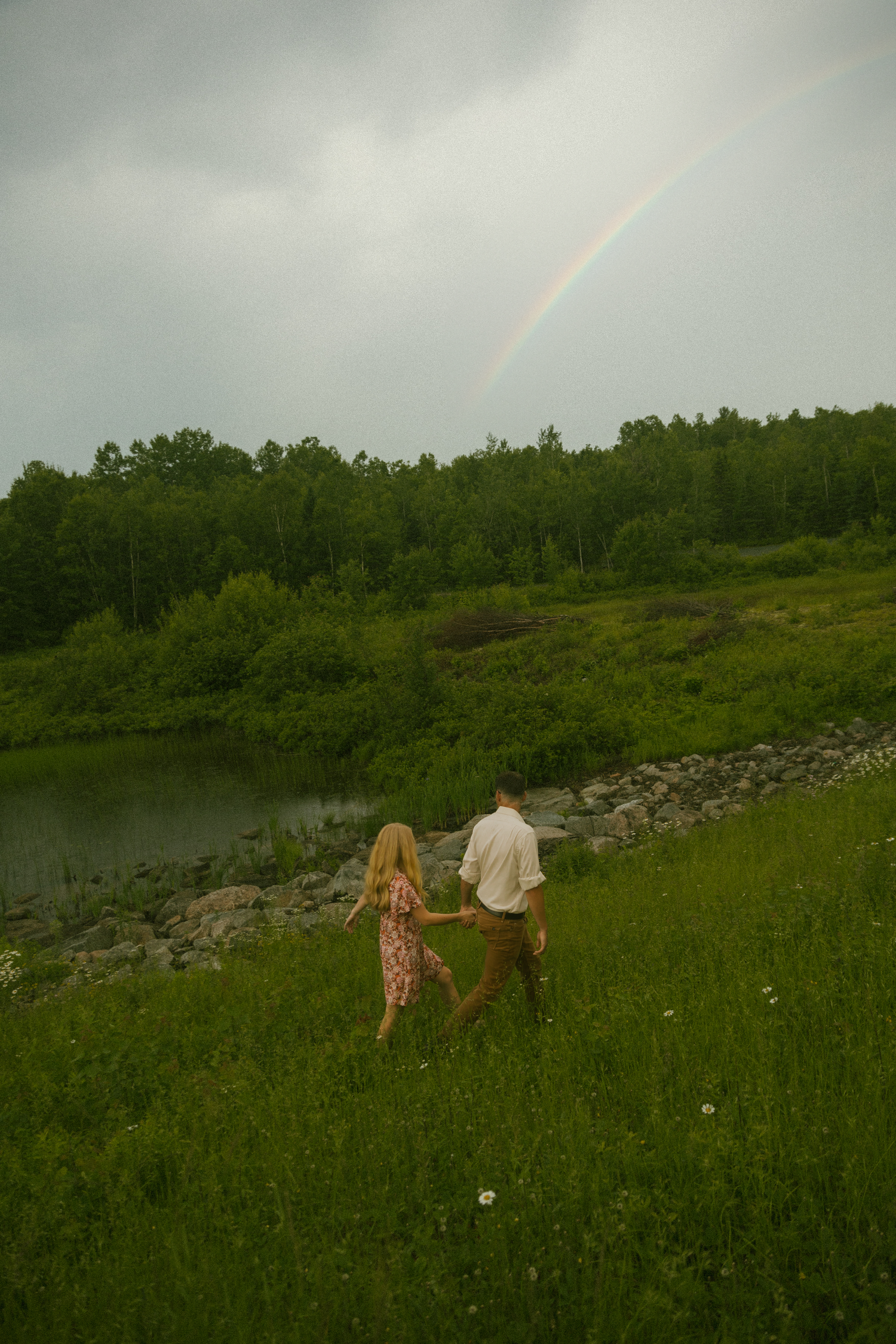 A couple walks through a lush green field with a rainbow visible in the cloudy sky.