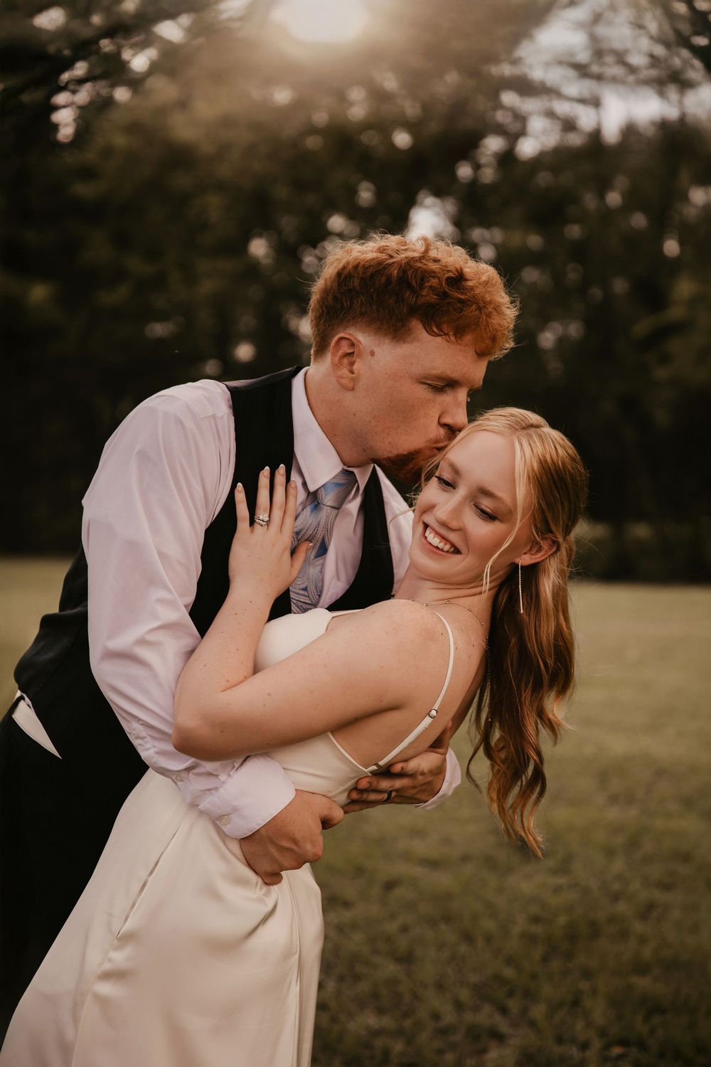 A romantic couple shares tender kisses and embraces during their wedding photoshoot in a grassy field at golden hour.