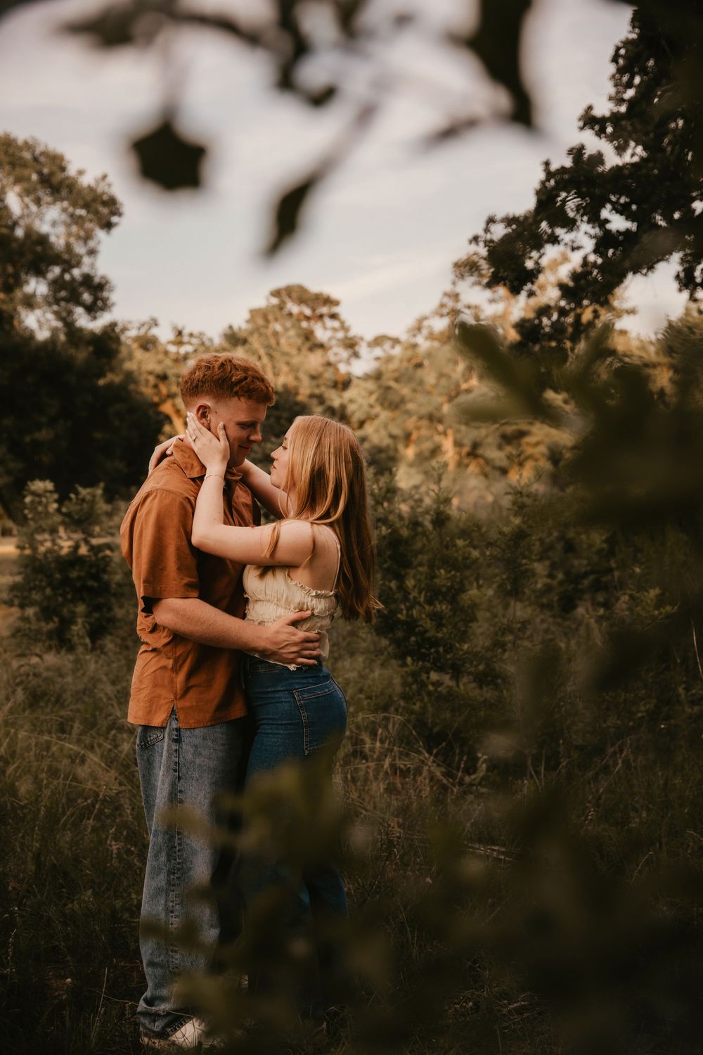 A couple shares an intimate embrace in a golden sunset-lit field with trees silhouetted in the background.