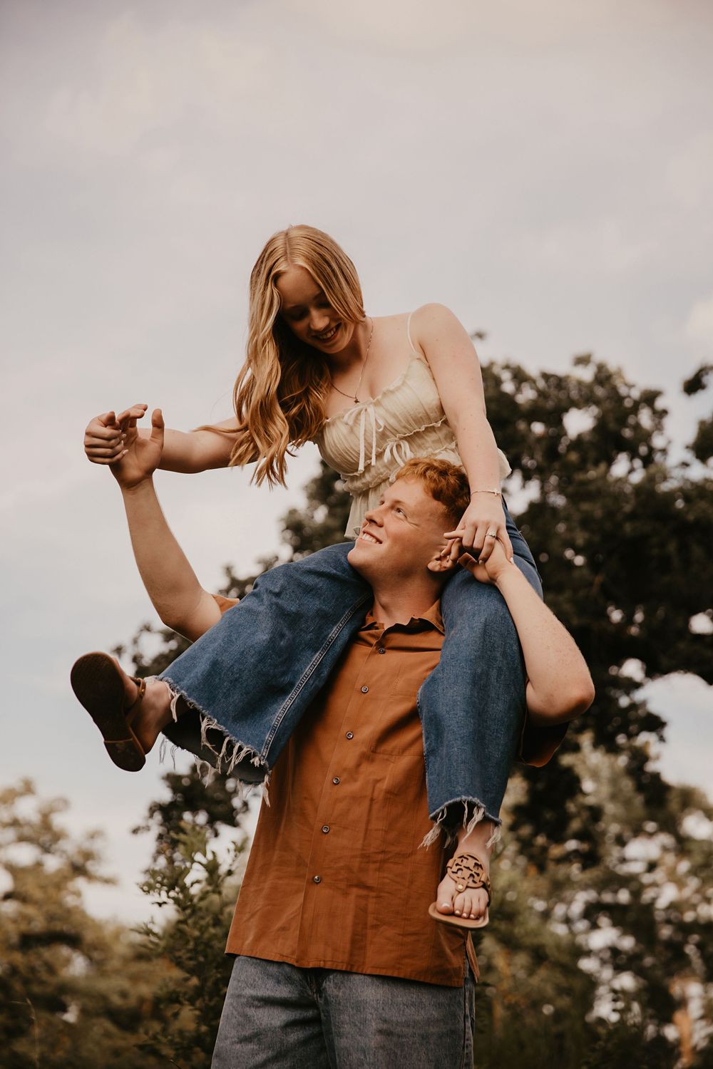 A couple sharing a playful moment outdoors as she sits on his shoulders in casual attire at sunset.