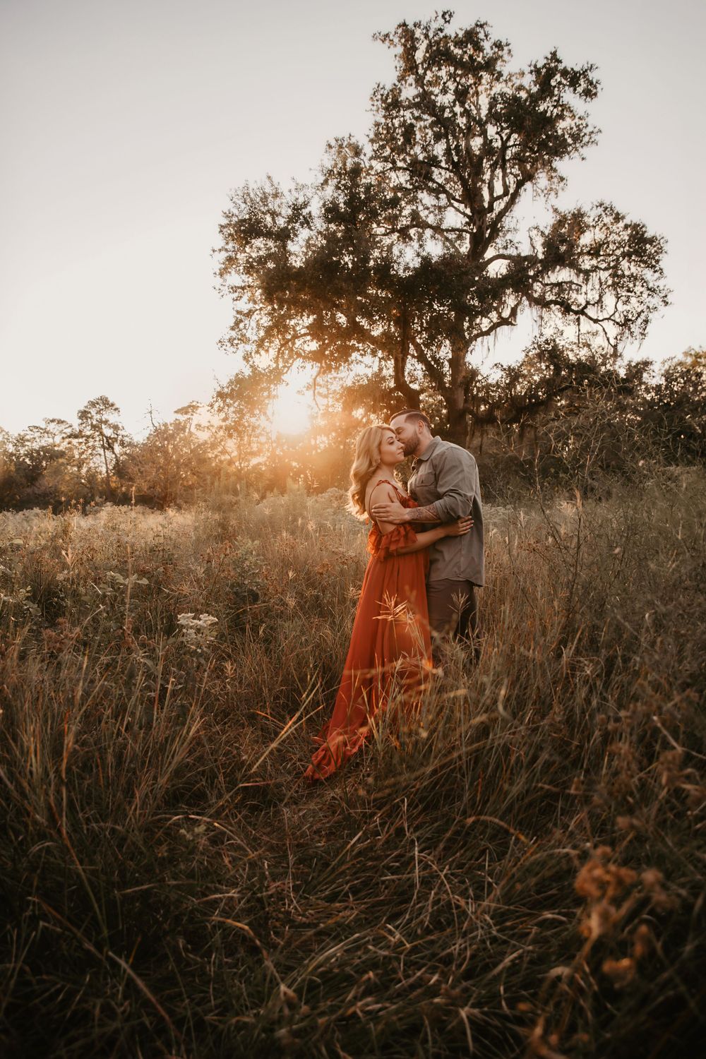 Romantic sunset portrait of couple embracing in tall grass beneath large oak tree.