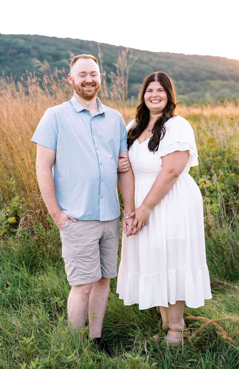 A couple poses together in a golden field during sunset wearing coordinated casual outfits.