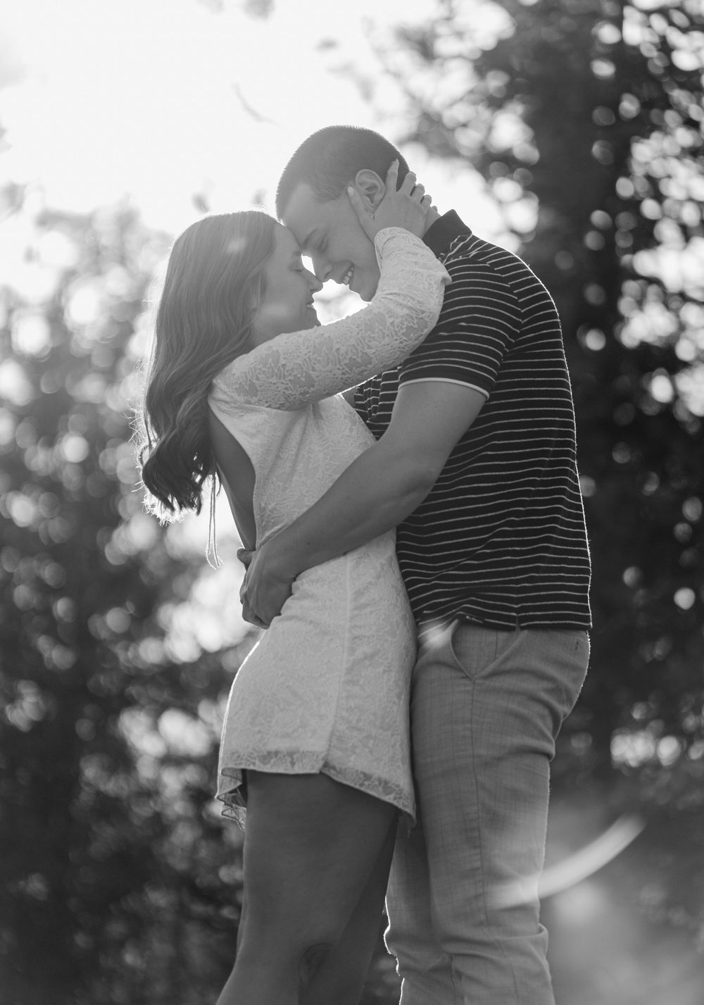 Black and white photo of a romantic couple embracing and kissing outdoors with soft bokeh trees in background.
