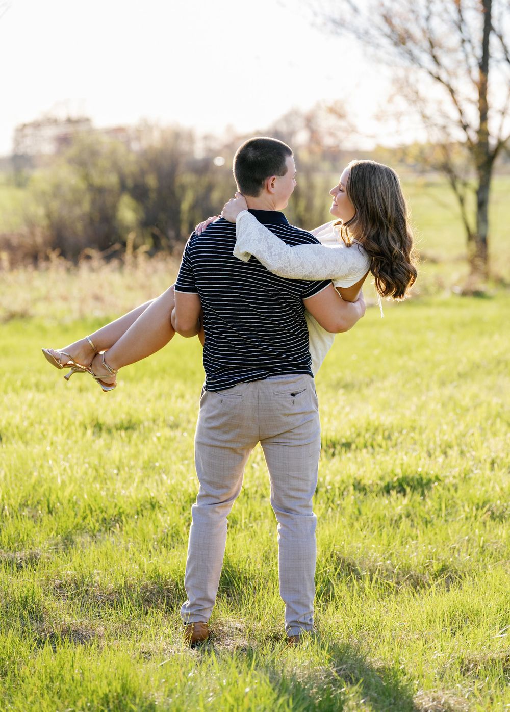 A man carries a woman in his arms while standing in a sunny green field during an outdoor engagement photo session.