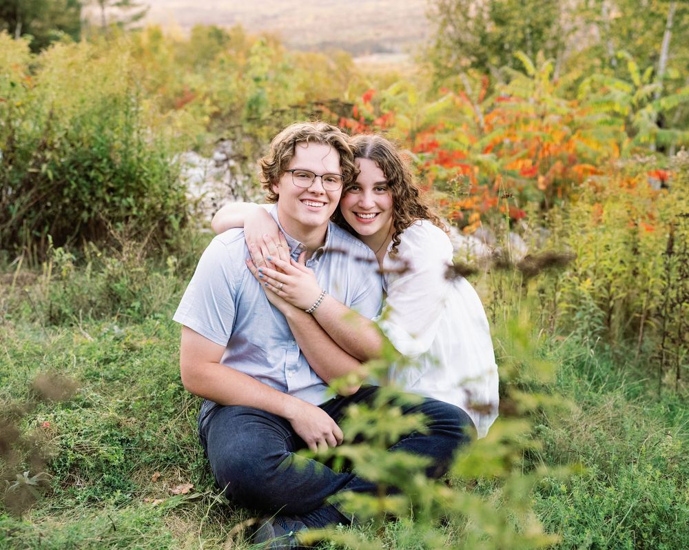 A couple poses together in a grassy field with autumn foliage in the background.