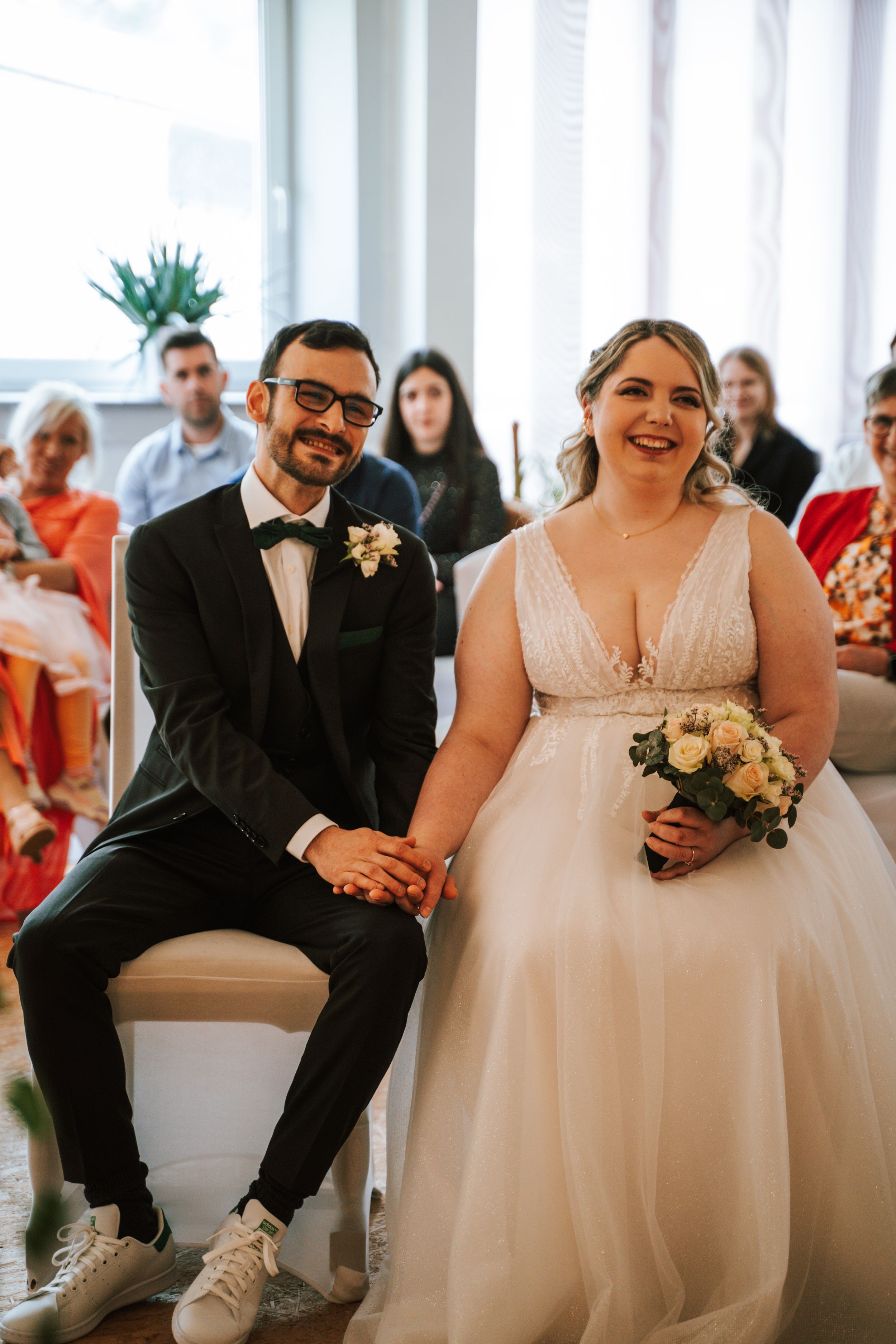 Wedding couple seated on white chairs with guests in background at bright indoor venue.