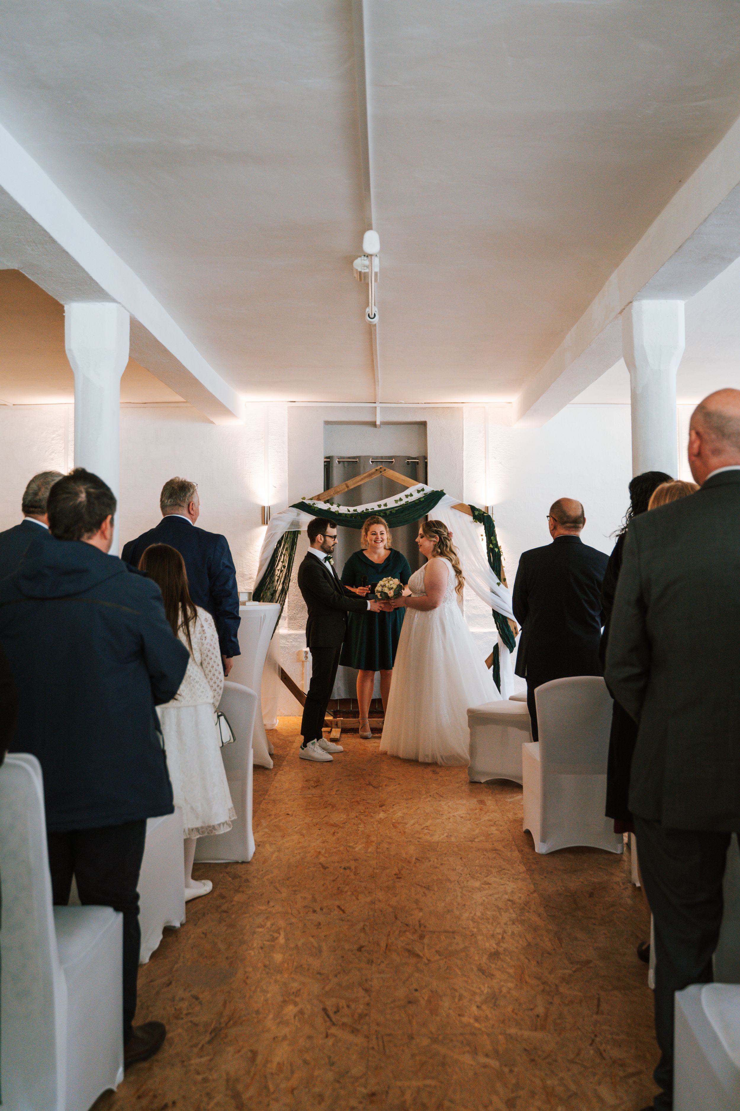Wedding guests gathered in a long white room with draped decorations and cork flooring.