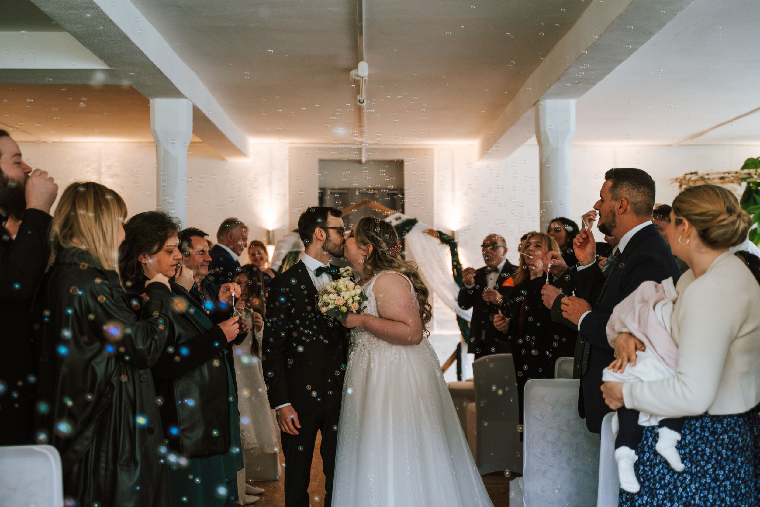 Wedding guests gathered indoors watching ceremony with joyful expressions.