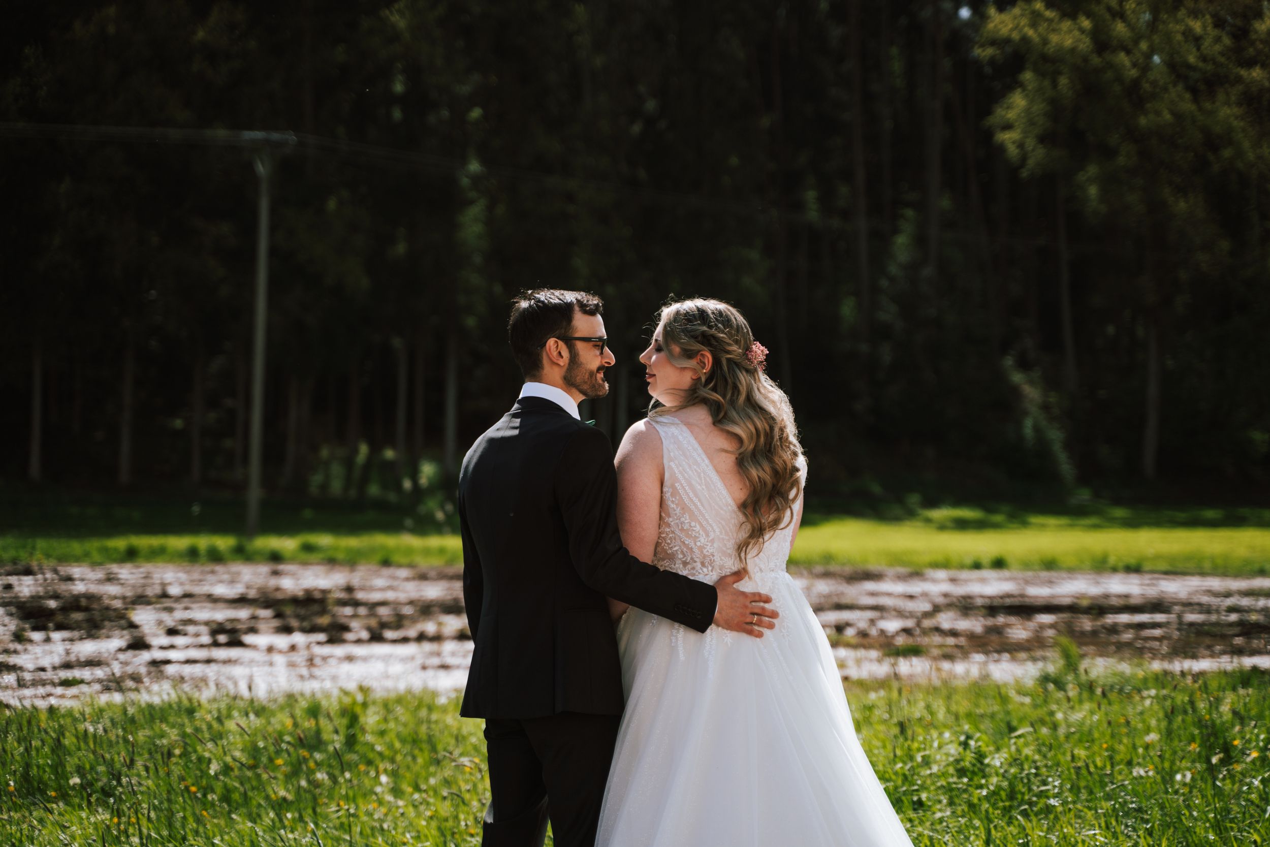 Newlyweds share an intimate moment in a sunlit field with forest backdrop.