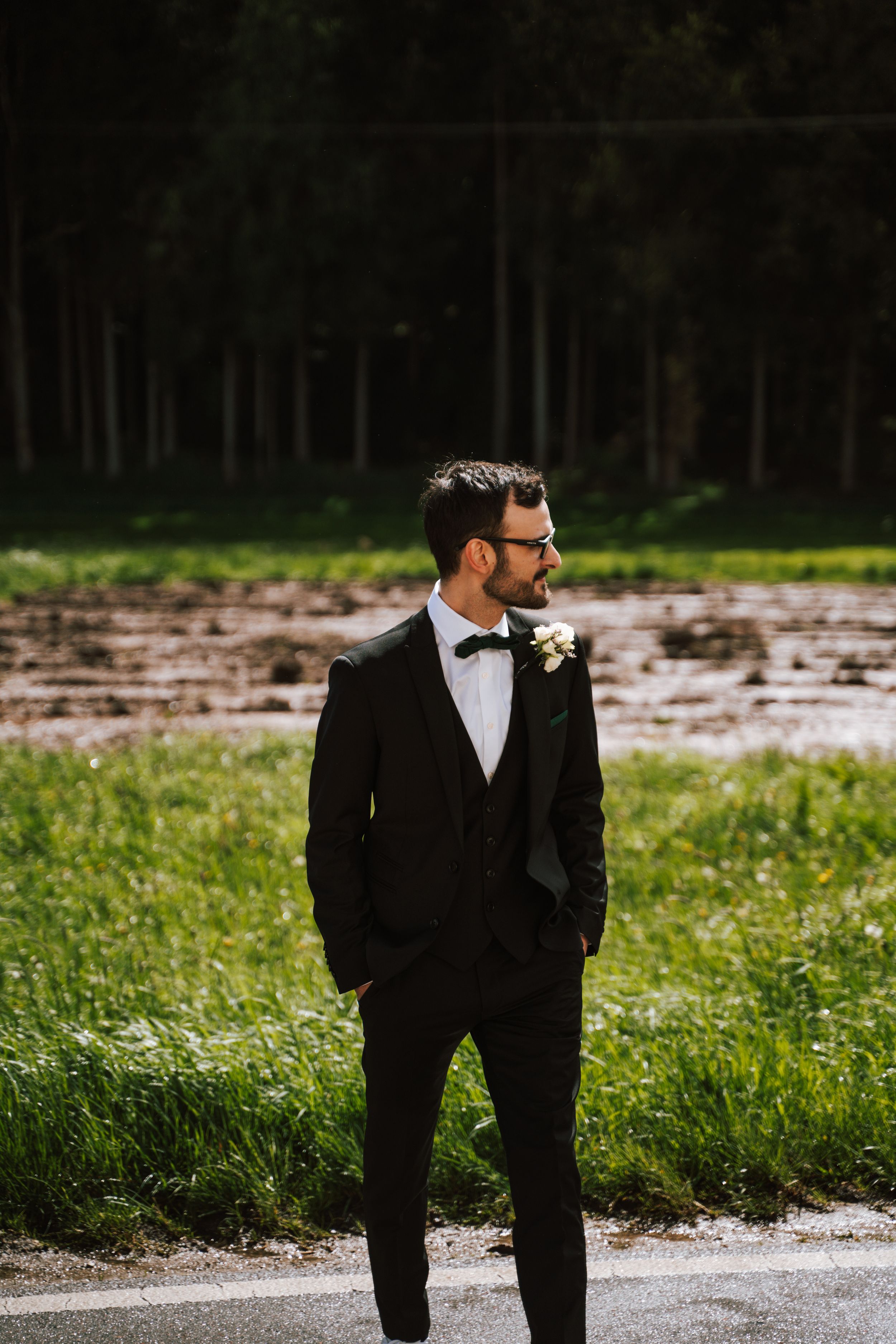 Groom in classic black tuxedo and bow tie poses in outdoor setting.