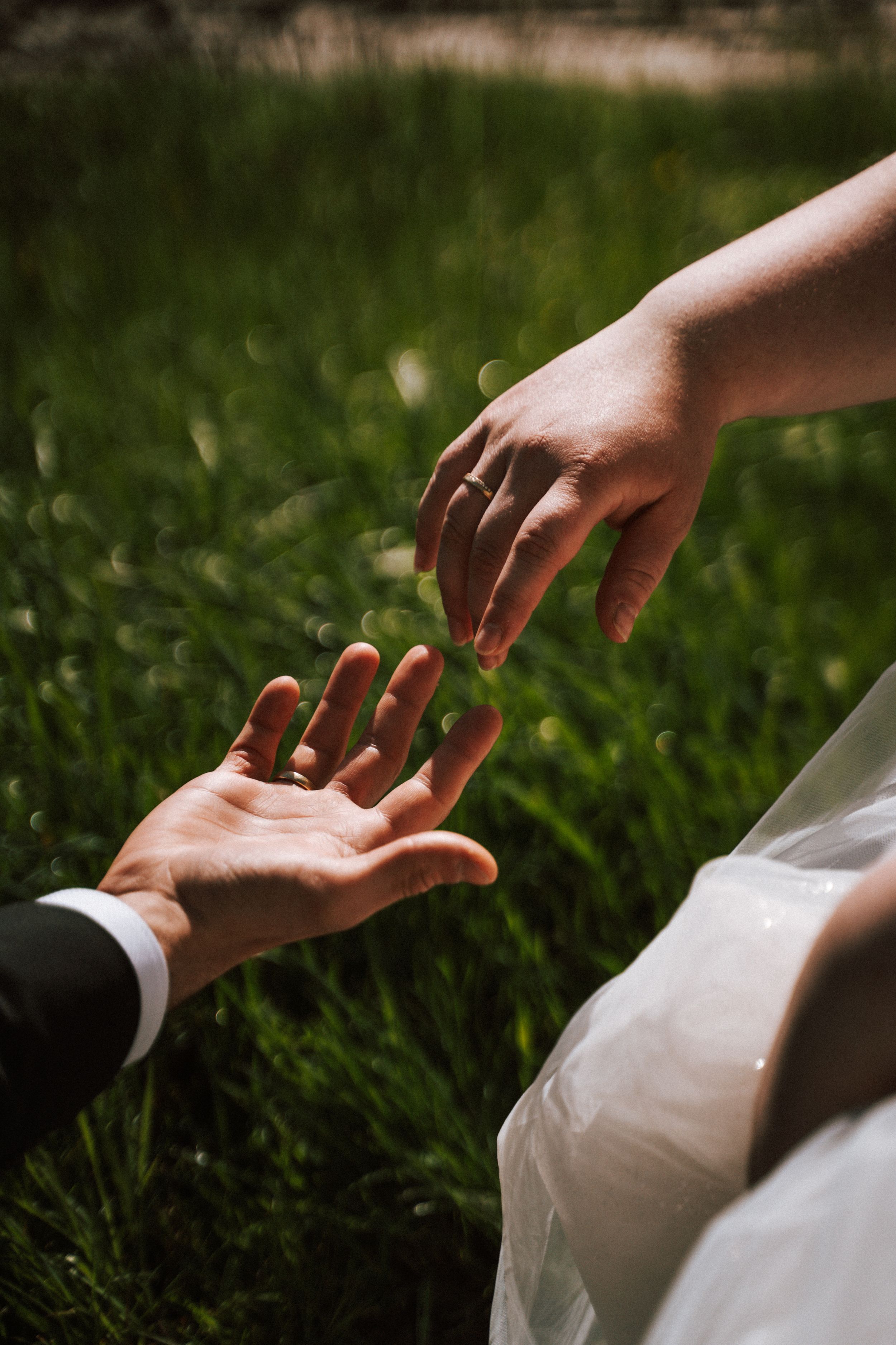 Close-up of wedding couple's hands reaching toward each other over grass.