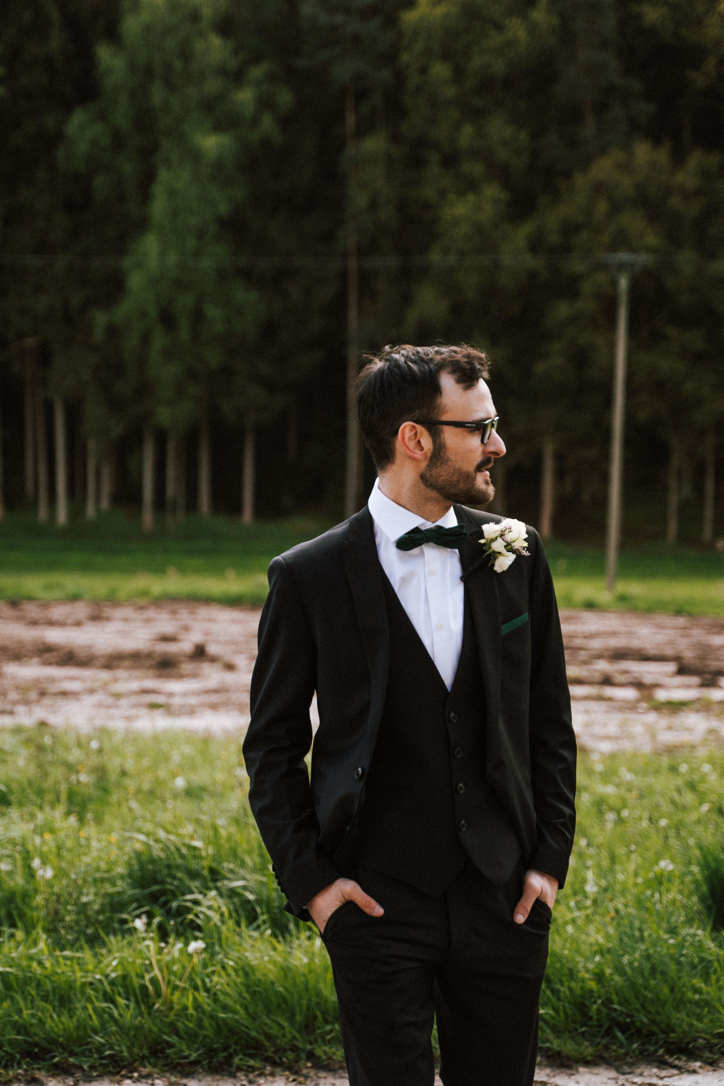 Groom in classic black tuxedo and glasses stands in outdoor setting with forest backdrop.