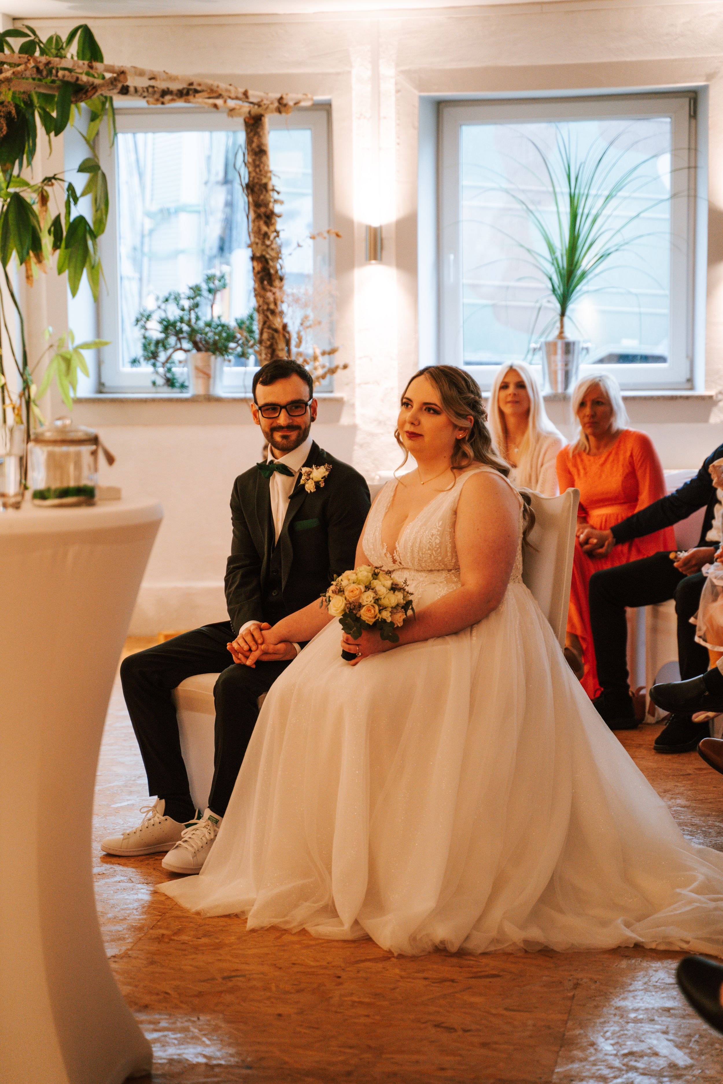 Wedding celebration in a bright indoor venue with white chairs and potted plants by the windows.