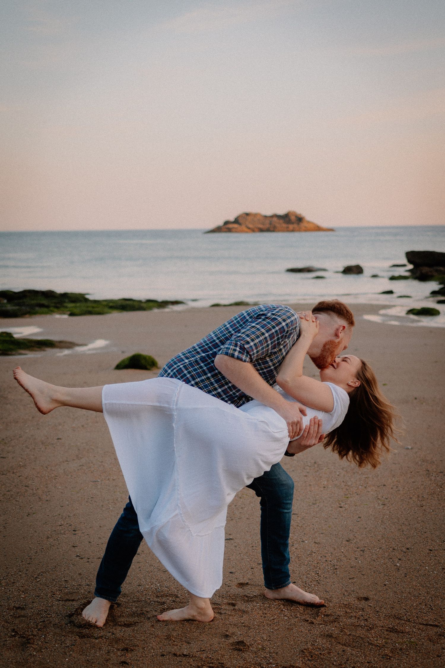 A romantic couple dips on the beach with an island visible in the distance at sunset.