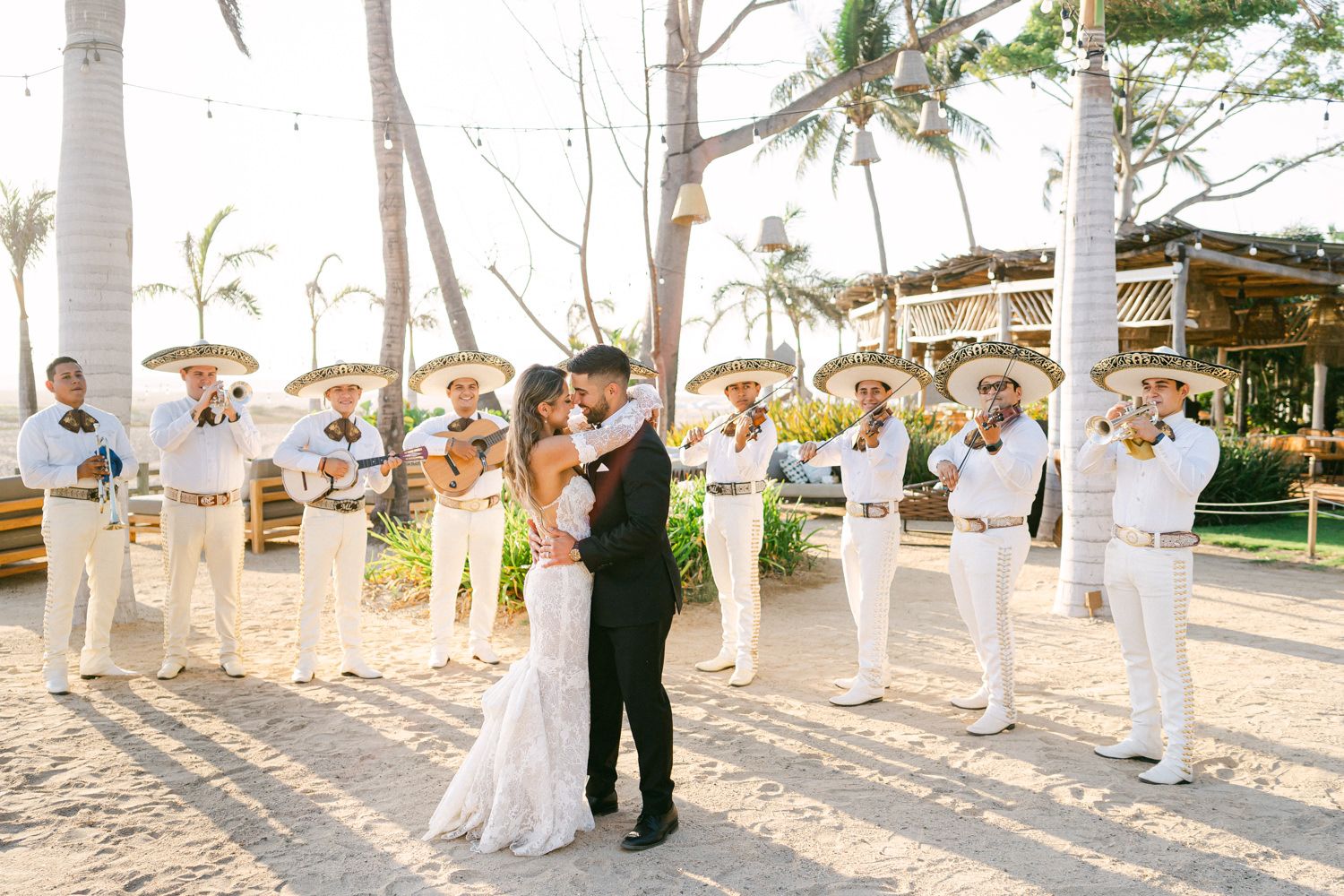 A wedding couple dances while guests in white attire form a circle around them on a beach.