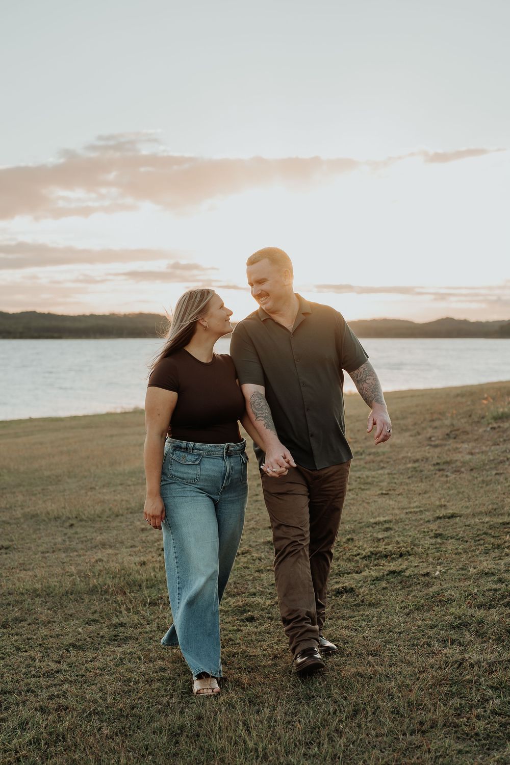 Couple holding hands walking by a lake at sunset, smiling at each other in casual outfits on green grass.