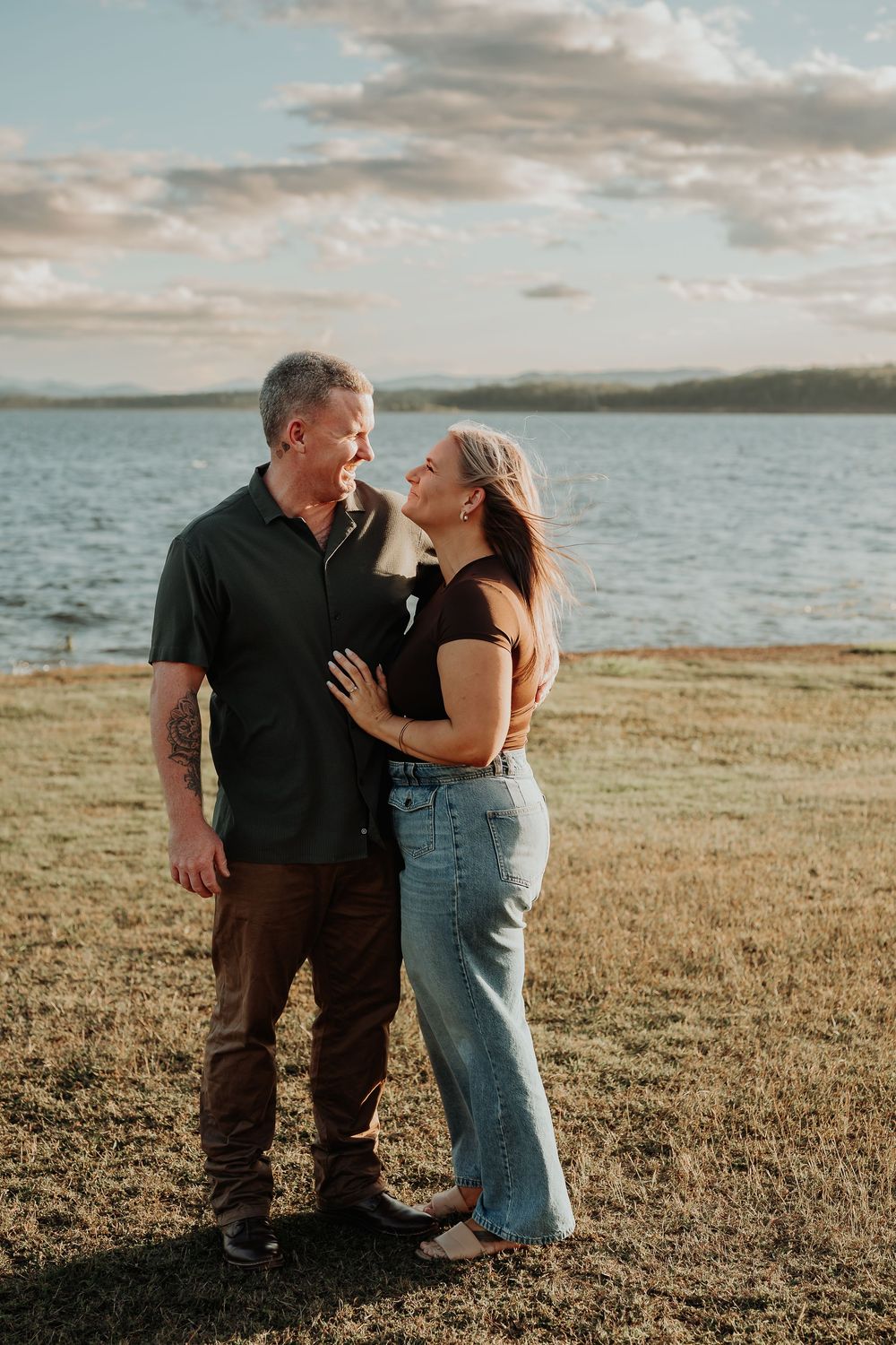 Couple sharing a romantic moment by a lake at golden hour, woman in jeans embracing man in dark shirt on green grass.