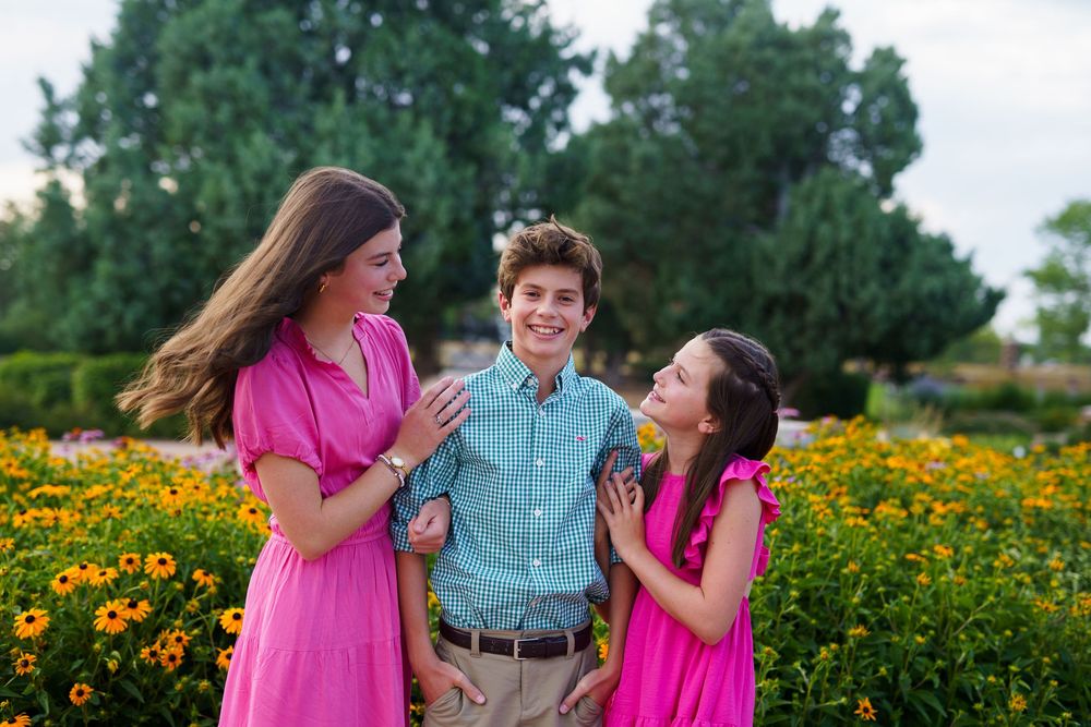 Three people in bright coordinated outfits laugh together in a flower garden.