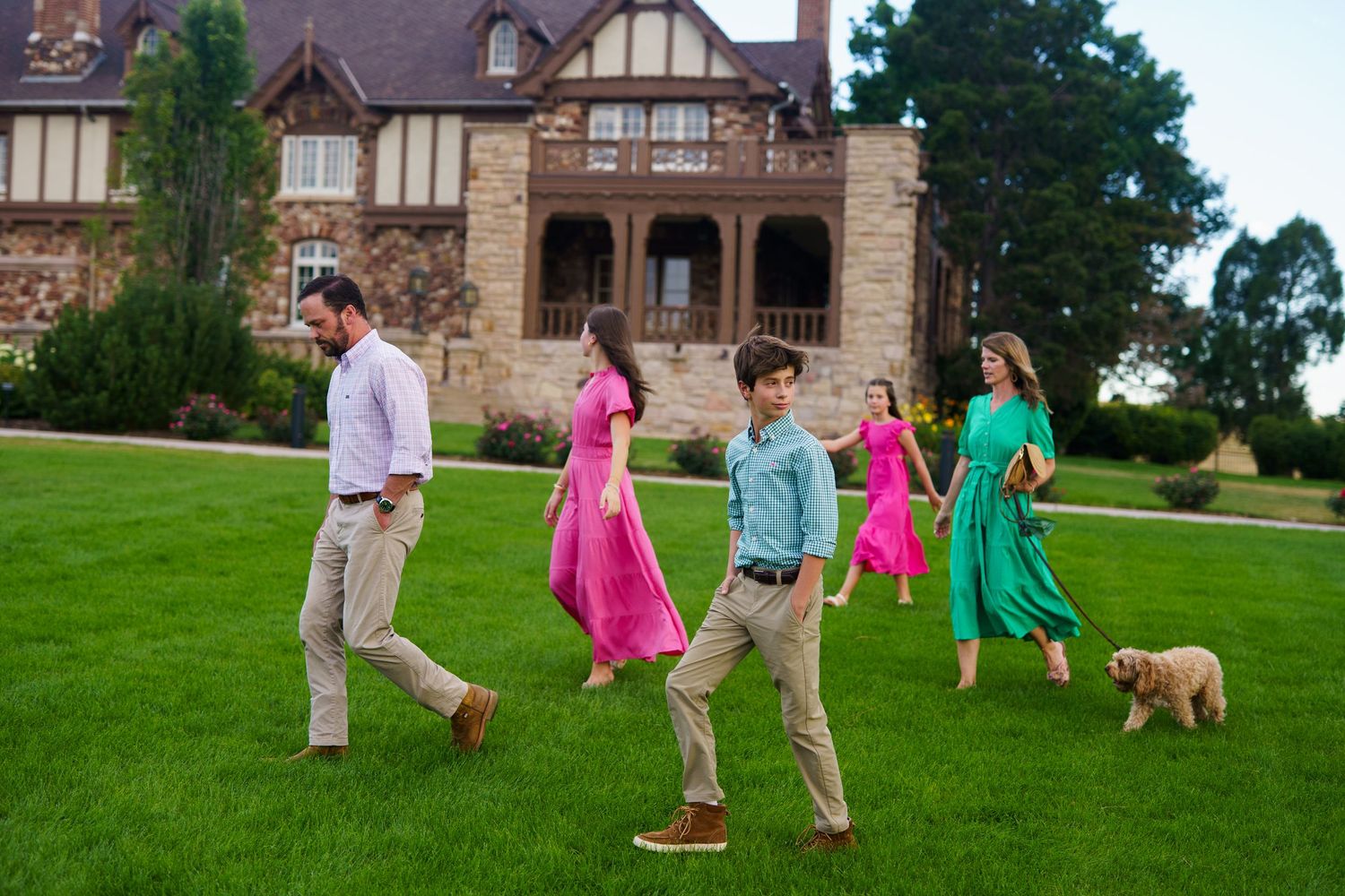 A family in coordinated outfits walks across a mansion lawn with their golden dog.