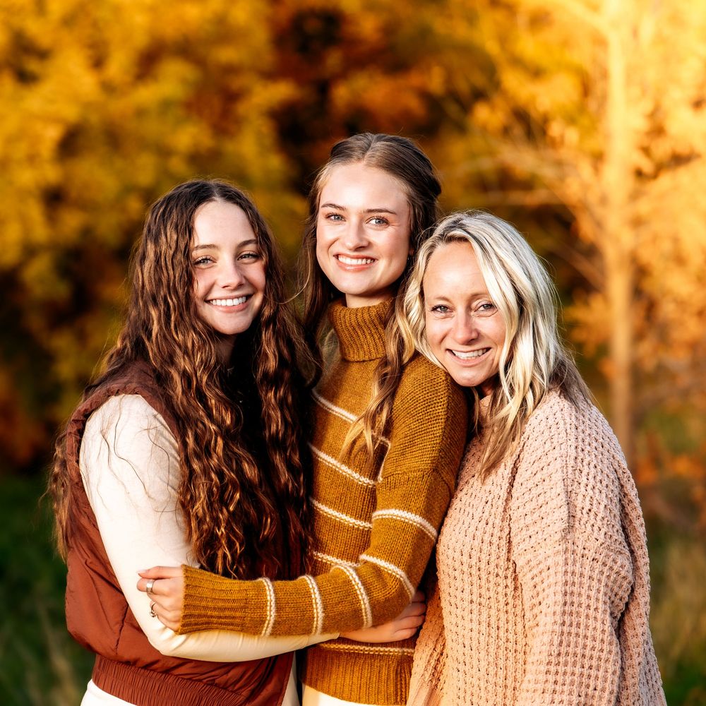 Three friends share joyful embraces while wearing cozy fall sweaters in a sunlit autumn setting with golden foliage.