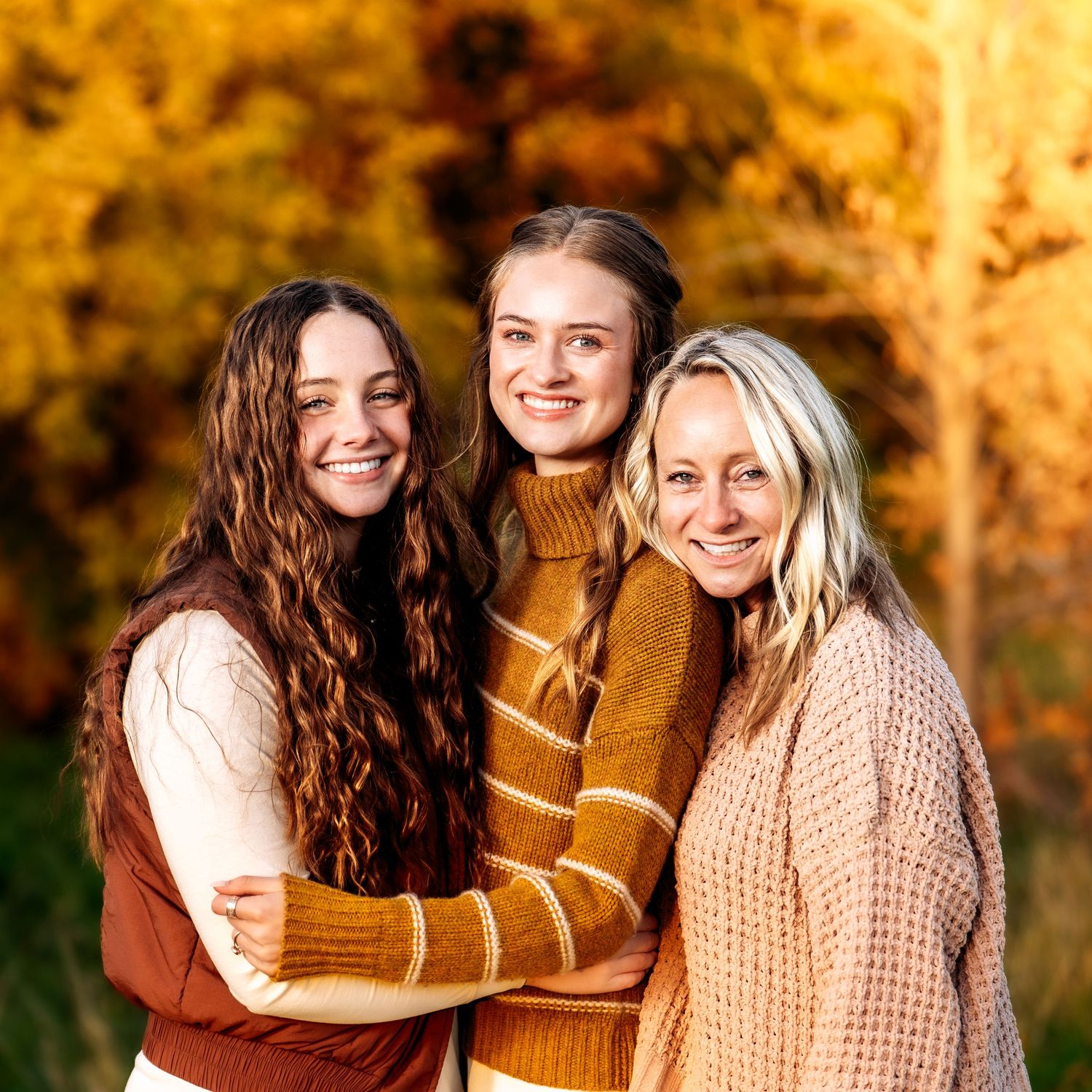 Three friends share joyful embraces while wearing cozy fall sweaters in a sunlit autumn setting with golden foliage.