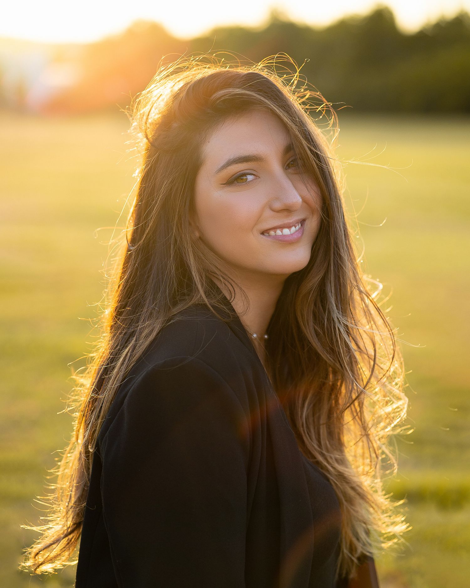 Young woman with long dark hair smiling over shoulder in golden sunset light in a green field.