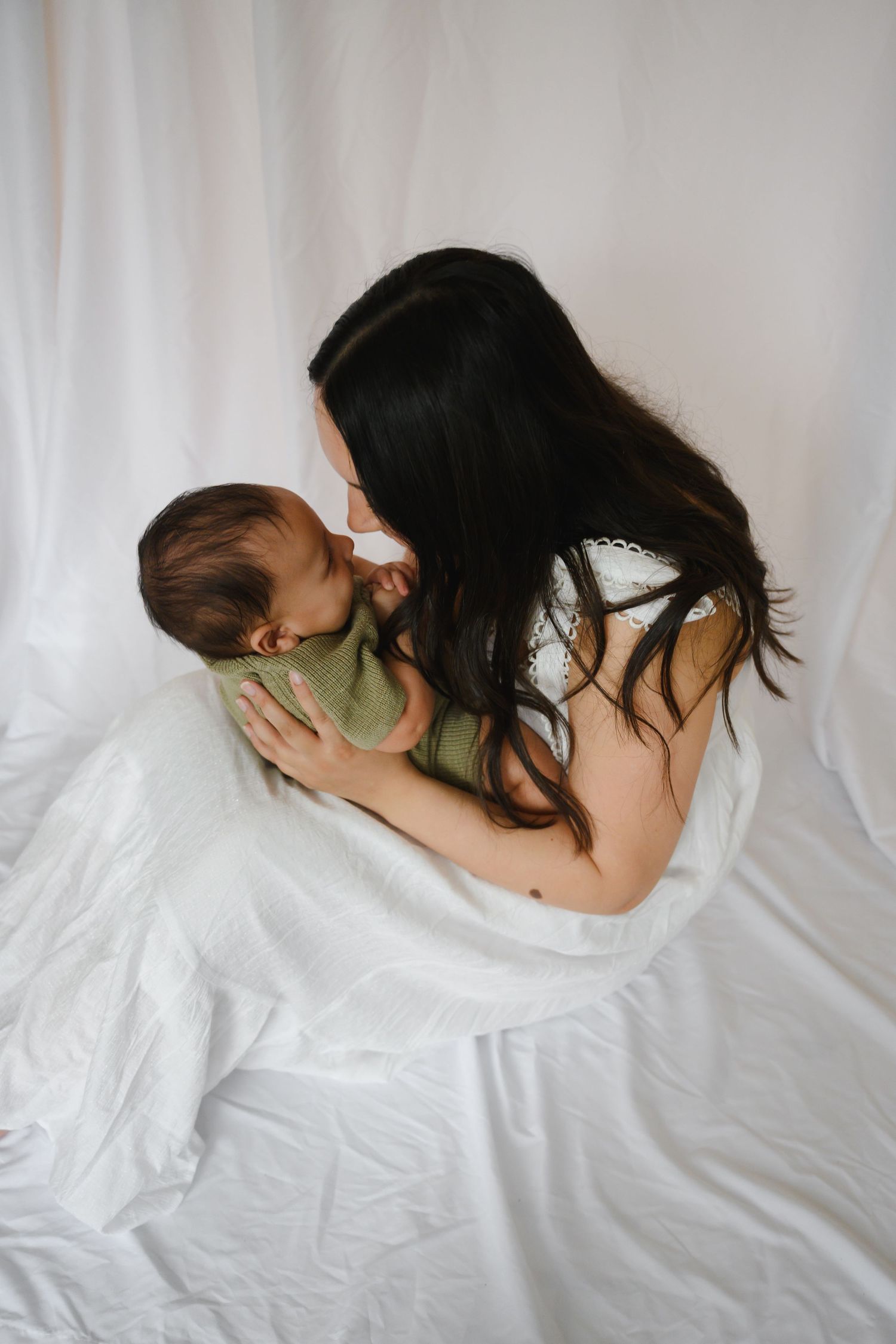 A dark-haired mother tenderly kisses her newborn baby wrapped in a green swaddle against a white backdrop.