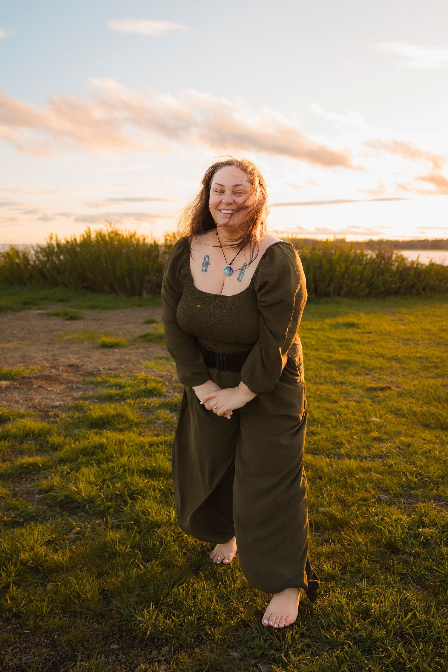 A barefoot person wearing a dark jumpsuit stands in a grassy field at sunset with golden light and water in the background.