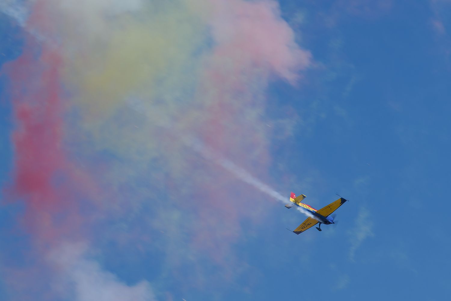Small aircraft flies through colorful smoke trails against blue sky.