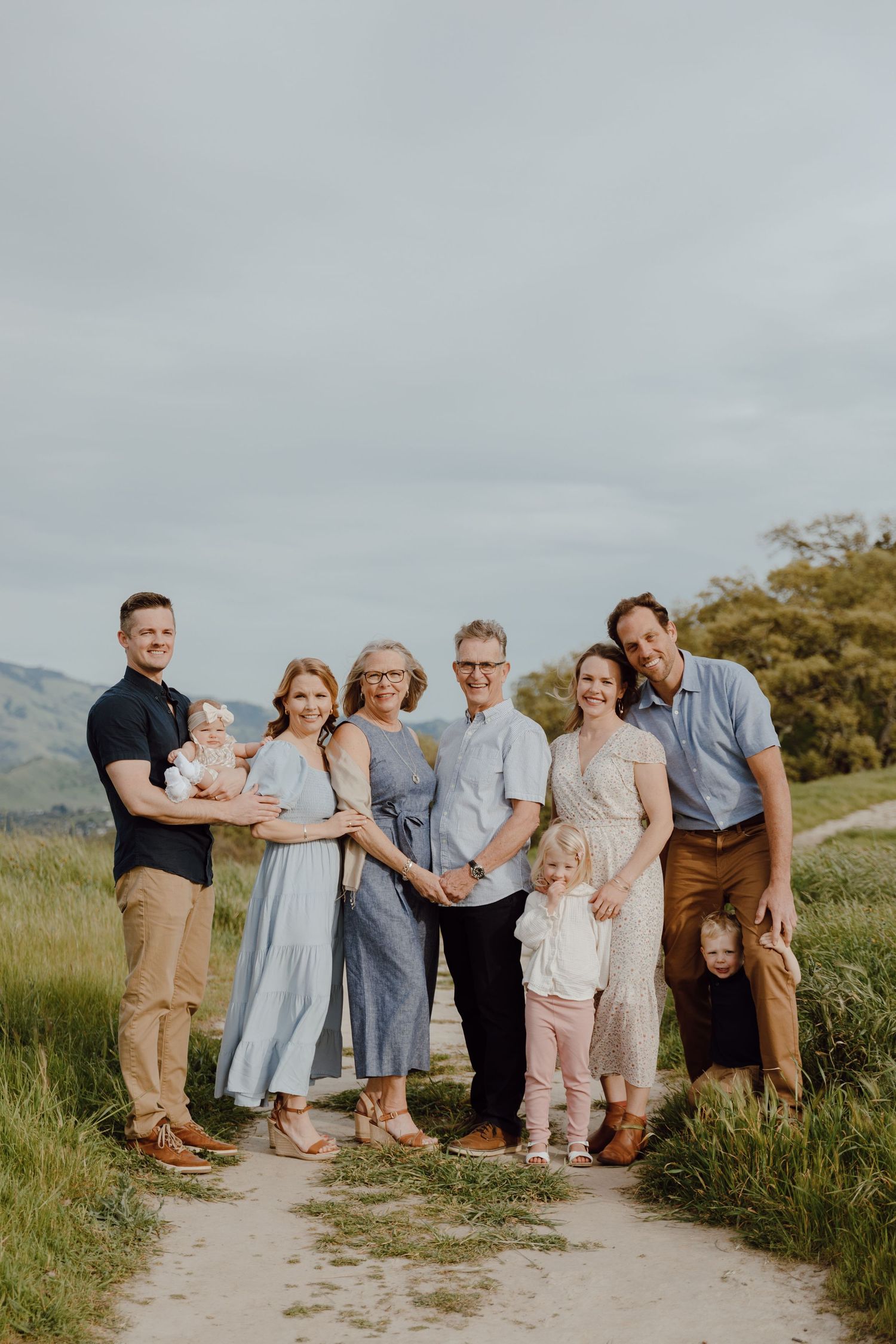 A multi-generational family poses together on a dirt path in a scenic mountainous landscape.