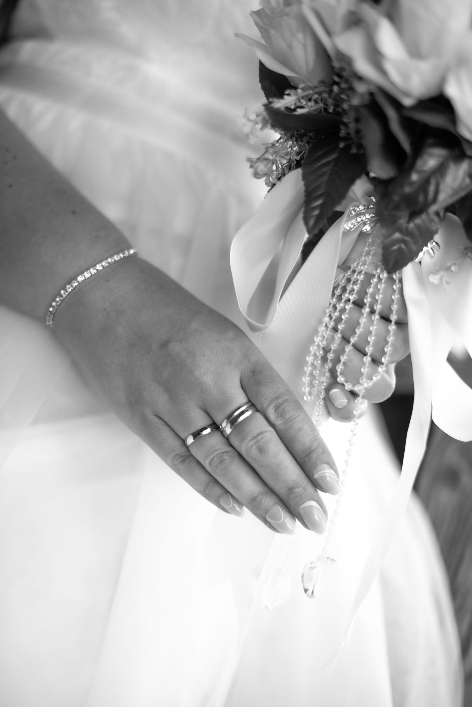 Close up black and white photo of a bride's hand with rings and pearl accessories against her dress.