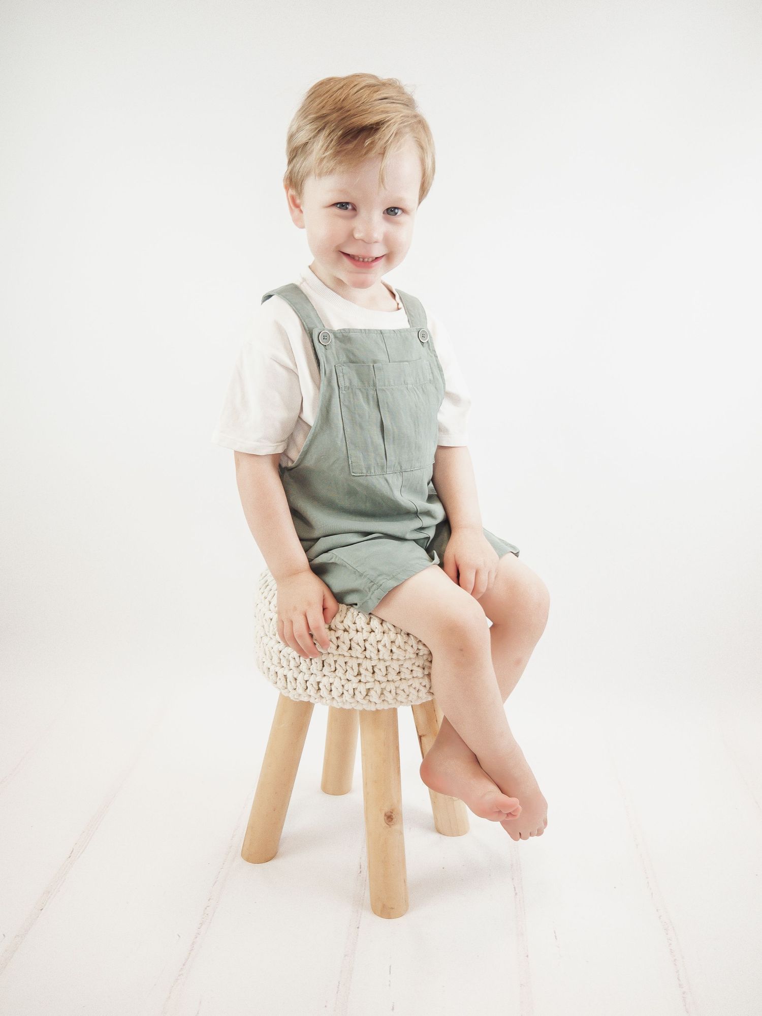 Small child in sage green overalls sits on a wooden stool with a woven seat against a white background.