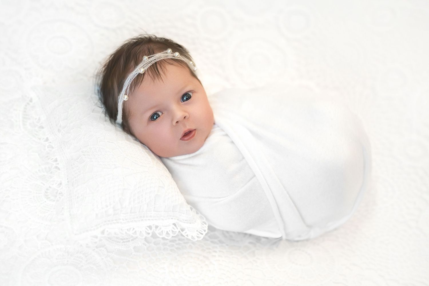 A baby wearing a white headband and swaddle lies on soft white fabric with delicate lace details.