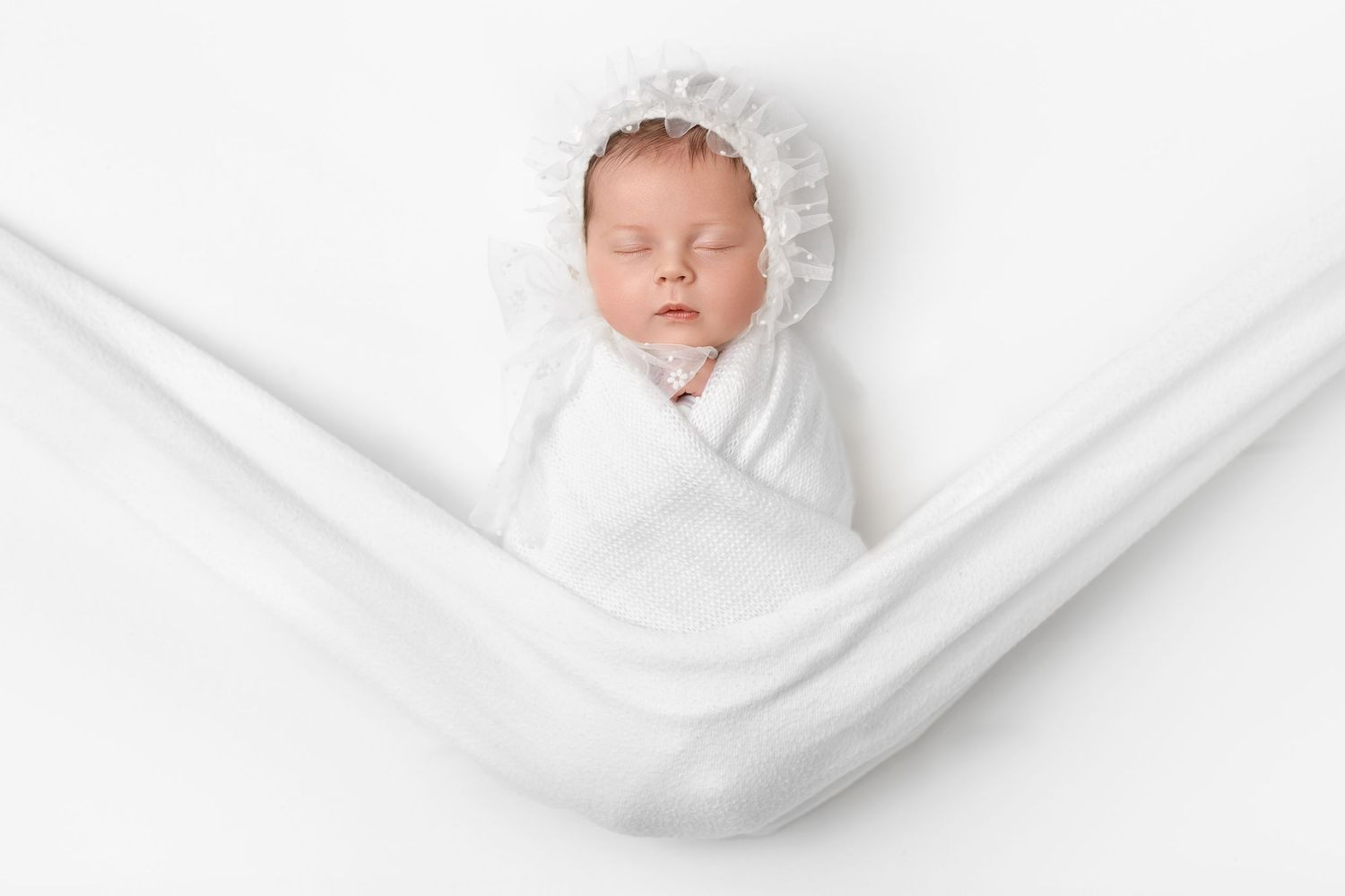 Newborn sleeping peacefully swaddled in soft white blanket and bonnet against white fabric background.