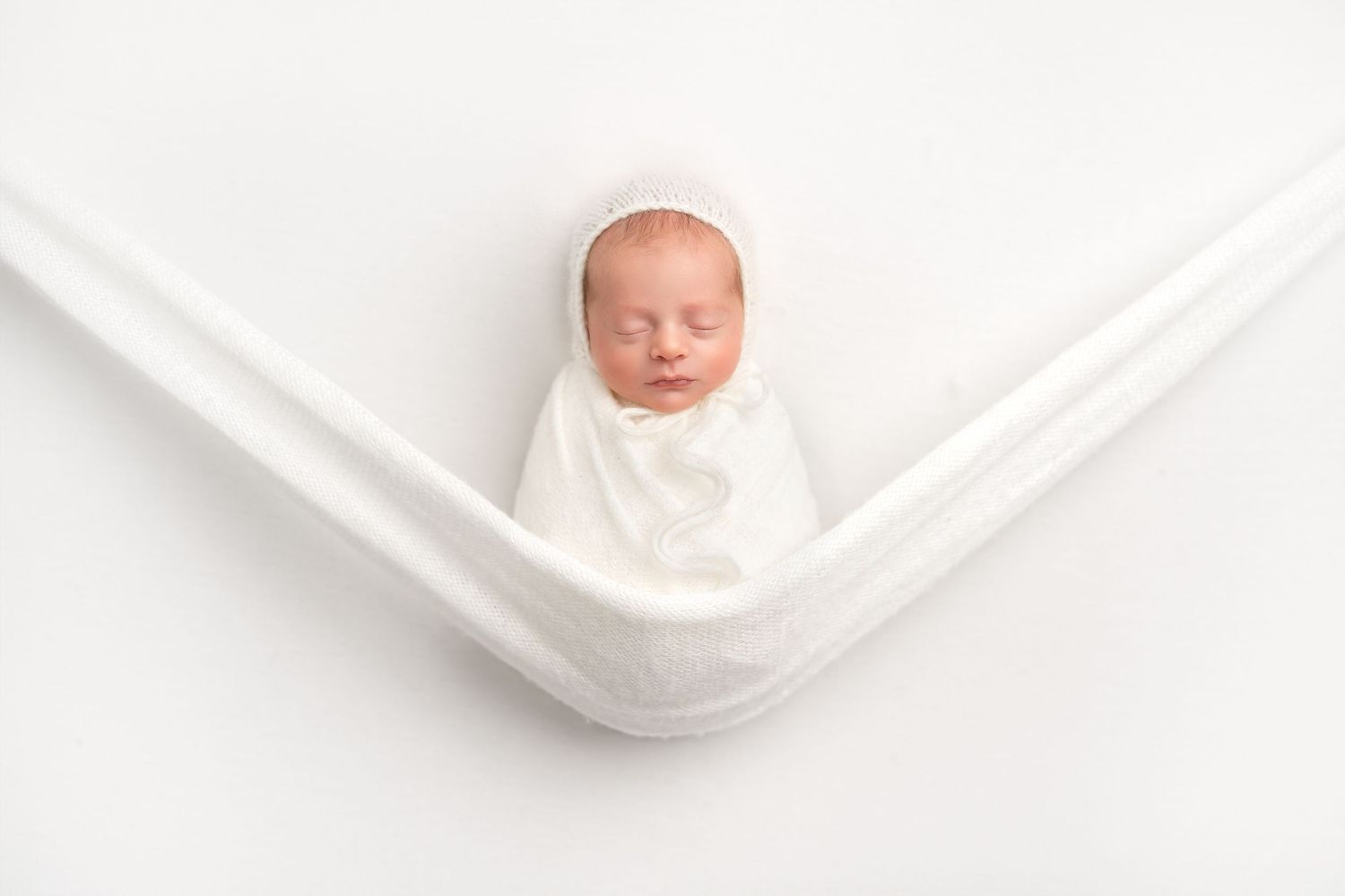 Newborn baby sleeping peacefully in a white hammock-style wrap against a light background.