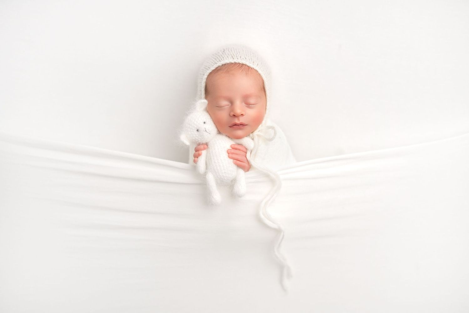 A sleeping newborn baby swaddled in white fabric lies peacefully against a soft white backdrop in a minimalist portrait.
