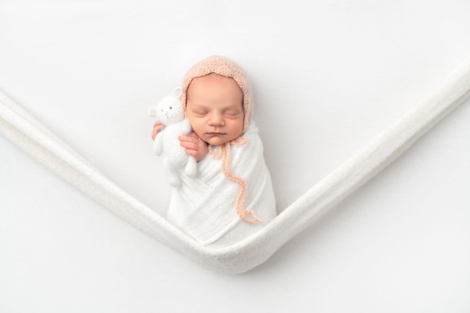 Sleeping newborn baby swaddled in white blanket nestles peacefully in curved white fabric backdrop.