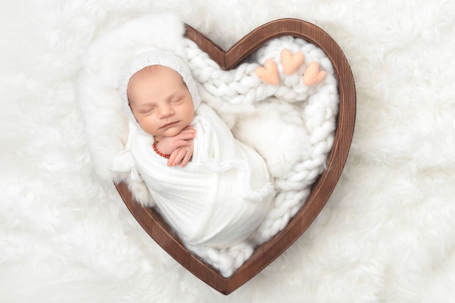 A sleeping newborn wrapped in white swaddle lies in a heart-shaped wooden container on soft white fabric.