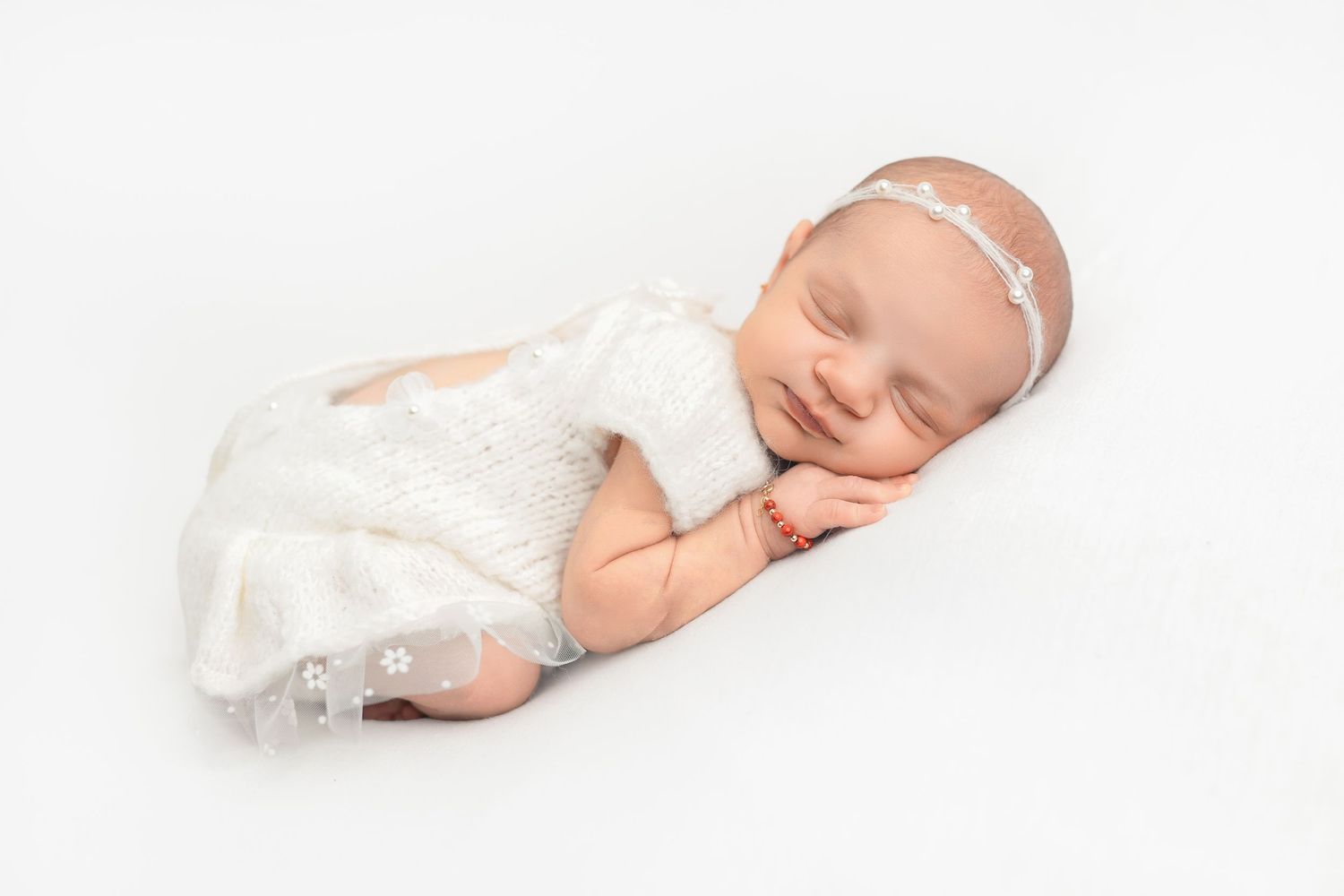 A sleeping newborn baby in soft white clothing lies peacefully on a white background.