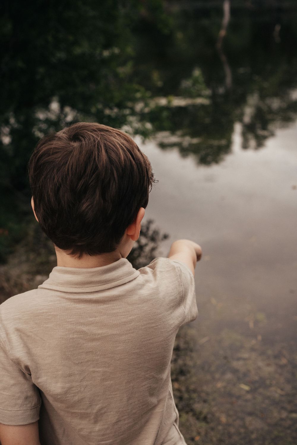 Child pointing at something across a serene lake while standing on the shore.