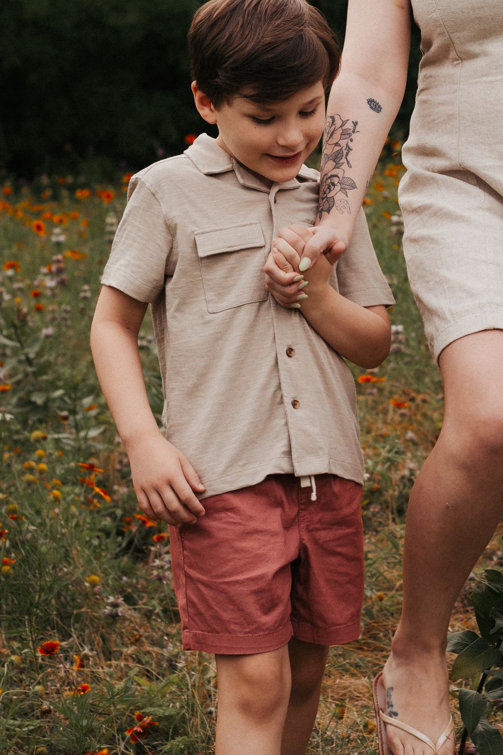 Child in beige shirt holding adult's tattooed arm in a field of orange wildflowers.