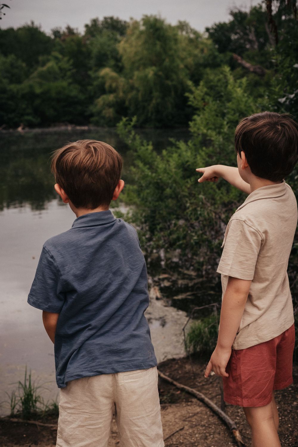 Two children standing by a lake pointing at something in the distance.