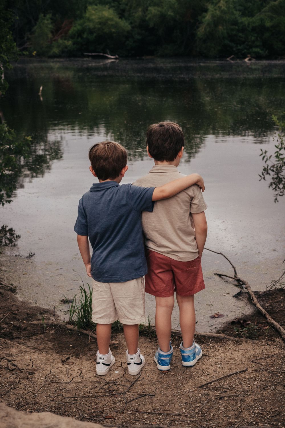Two young siblings standing arm in arm at the edge of a peaceful lake.