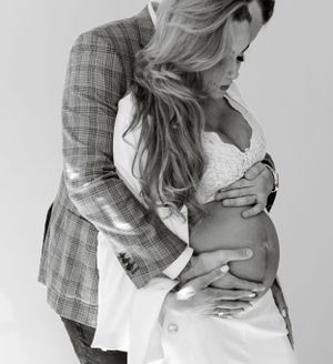 Black and white maternity photo showing a pregnant belly with hands wearing a plaid blazer and white dress.