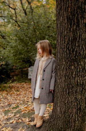 A figure in a plaid coat and tan boots stands beside a large tree trunk in an autumn setting.