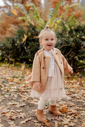 Small child in beige coat and boots plays in fallen autumn leaves.