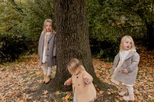 Three children in coordinating coats play around a large tree trunk.