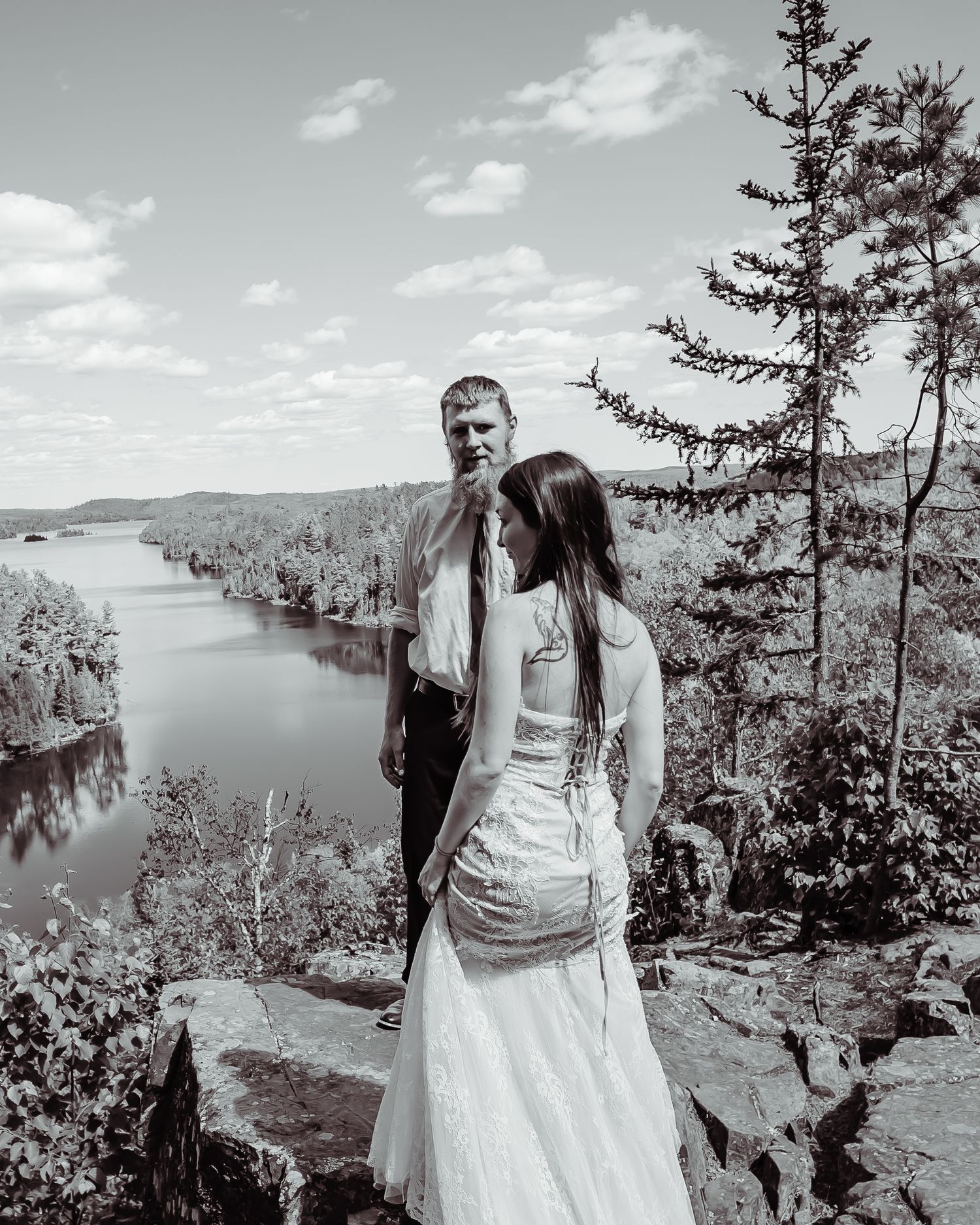 Couple in wedding attire standing on cliff edge overlooking a scenic lake vista in black and white.